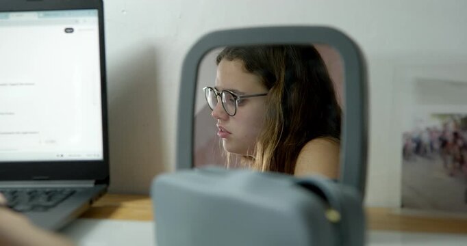 Student Stress Reflected: A young student stares into a small mirror, her reflection revealing a look of stress and concentration as she works on her laptop.