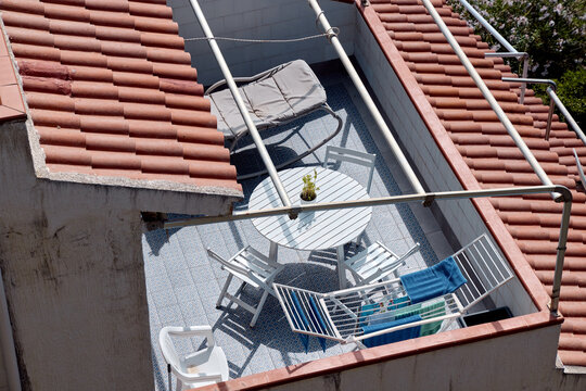 High angle view of a private Mediterranean rooftop terrace with a round table, chairs, sun lounger, and clothes drying rack between tile roofs.