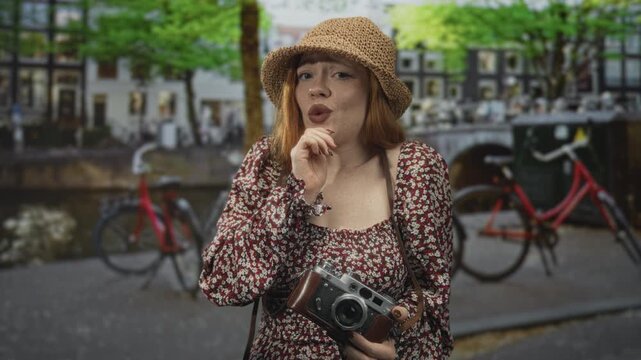 Woman redhead in straw hat holding retro camera with finger to chin thinking on a canal street with bicycles and buildings visible; curiosity wonder.