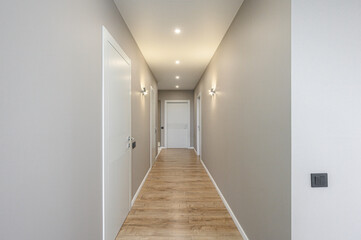 Fototapeta premium narrow modern hallway with light grey walls, warm wooden laminate flooring, white interior doors, recessed ceiling lights, and stylish wall sconces; minimalist design