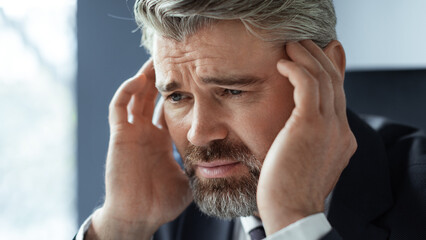 Fototapeta premium Worried Middle Aged Caucasian Man Holding Head In Distress While Sitting At Desk In Office. Struggling With Stress And Work Pressure, Seeking Calmness And Focus To Regain Mental Clarity