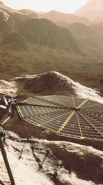 A solar panel array sits on rocky terrain in a barren landscape. Sunlight shines on the panels while distant mountains create a backdrop. The scene appears to be on another planet.