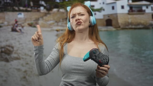 Young woman wearing cat ear headphones holds gamepad and points finger in front of seaside building while pouting and squinting in displeasure; frustration defiance.