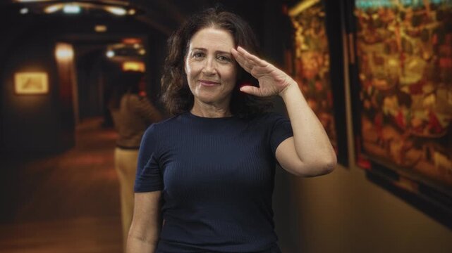 Middle age woman salutes with right hand to forehead beside framed painting in gallery building wearing navy shirt; contemplation memory.