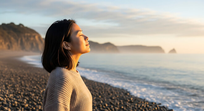 Woman stands on rocky beach looking out at calm ocean waves at sunset