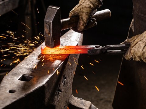A blacksmith strikes a glowing piece of hot metal on an anvil with a hammer, producing bright sparks.