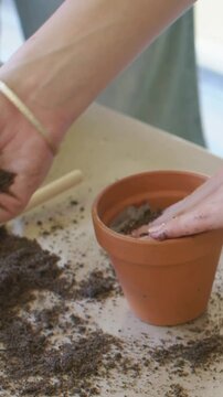 Vertical video: Scooping hands moving soil from pile packing into pot on table to plant with dowel