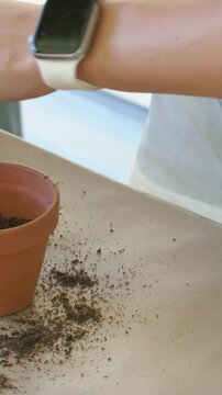 Vertical video: Scooping soil pile, arm wearing white watch, top filling clay pot to plant on table
