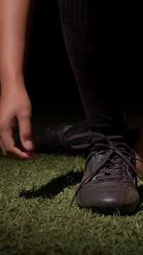 Vertical video: Reaching player kneeling tying black shoelaces on spotlit turf for sport in socks