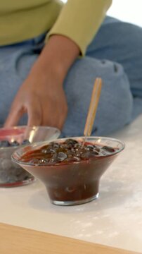 Vertical video: Right hand entering man in jeans, green top preparing red bowl beside dessert bowl