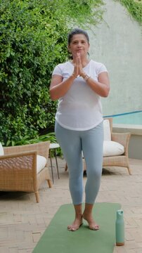 Vertical video: Breathing woman shifting, balancing on green mat on patio by pool, in top, bottle