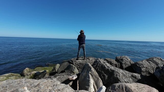 Tranquil seaside scene featuring a solitary angler fishing from rugged rocks with an endless ocean horizon and peaceful weather conditions.