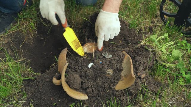 Individual using a small trowel to excavate soil around a broken pot and coins in a grassy area, revealing hidden treasures during a metal detecting activity