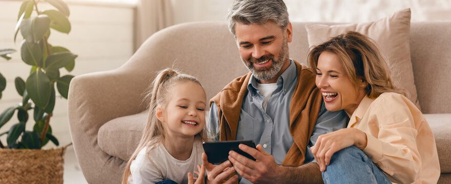 Happy Family Of Two Adults And One Child Smiling Together As They Watch Something On The Mobile Device, Sitting On A Couch In A Cozy Living Room, Enjoying Family Bonding Time
