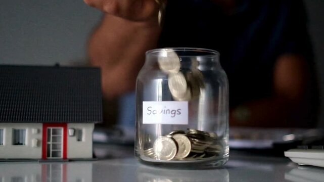 Hand Dropping Coins into Savings Jar Next to Model House for Real Estate Investment