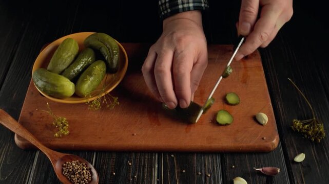 Hands slicing pickles on a wooden cutting board with a bowl of whole pickles and spices visible in a cozy kitchen setting
