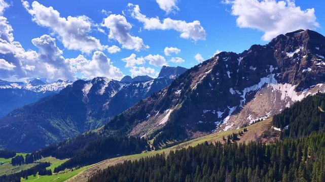Forward drone flight above alpine mountains, forests and remaining spring snowfields under dramatic clouds in the French Alps