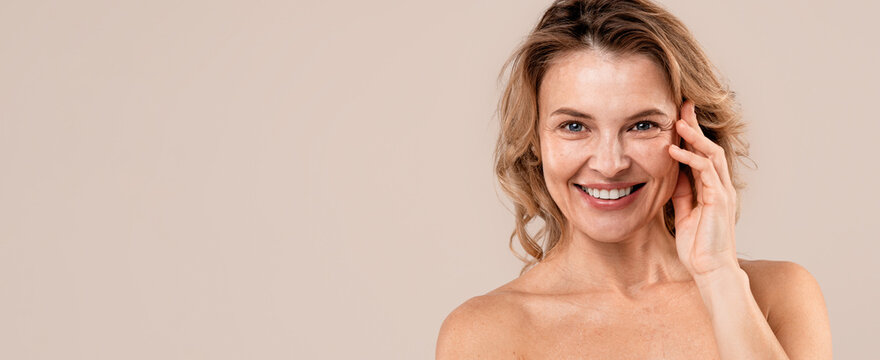 Cheerful Middle Aged Woman Smiling And Touching Her Face, Showcasing Natural Beauty In A Studio Setting. Engaging In Skincare Routine, Focused On Self-care And Beauty