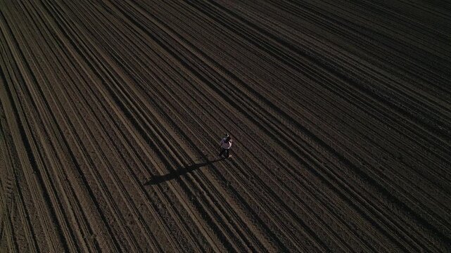 Aerial drone view flying over a lone person standing in the middle of a large, freshly plowed agricultural field with long, straight furrows, creating a sense of solitude and scale