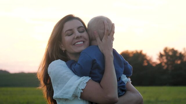 Tender moment of young mother gently hugging adorable baby boy while standing in greenfield during warm golden sunset. Showcasing maternal love and affection.