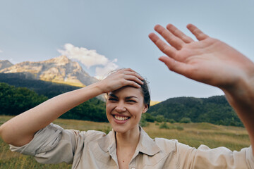 Naklejka premium Smiling woman in a light shirt raises her hand in a sunny open field, enjoying a mountain backdrop and the carefree mood of a rural day.