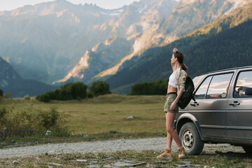 Naklejka premium Young woman with backpack stands beside a car in a mountain valley, taking a moment to soak in the rugged scenery during a daytime road trip