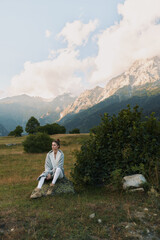 Naklejka premium Woman lifestyle sitting on rock in mountain meadow, casual outfit, thoughtful expression, natural light, scenic landscape, cloudy sky, peaceful atmosphere