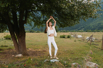 Naklejka premium Young woman in white croptop and pants dances under a large tree in a countryside field, radiating carefree joy and natural serenity.