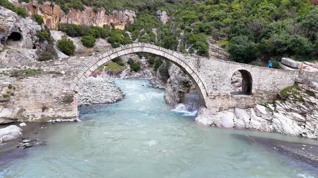 Aerial drone camera tracking left across the historic Ottoman-era Kadiut Bridge (Ura e Kadiut) spanning the turquoise Lengarica River canyon in Benje, Permet, Albania