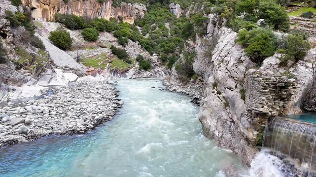 Aerial tracking shot pulling back underneath the grand stone arch of the historic Ura e Kadiut bridge, framing the rushing teal waters of the Benja River and the rugged Langarica Canyon