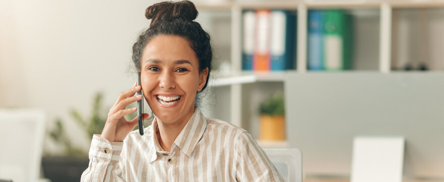Smiling Young Latina Woman Engaged In Phone Conversation At Office, Radiating Joy While Working In Professional Environment. Focused On Communication And Business Activities