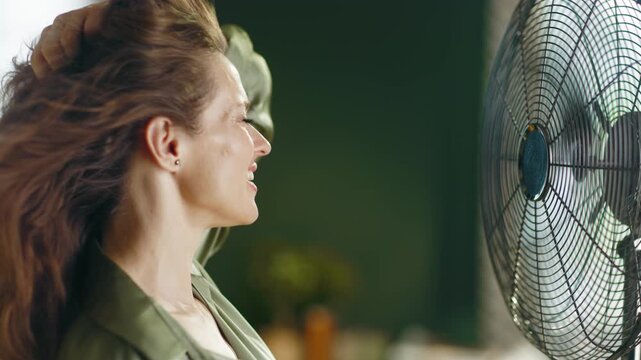 A happy woman with long brown hair, wearing a green shirt, experiences refreshing relief from summer heat as an electric fan blows her hair, conveying comfort and wellbeing.