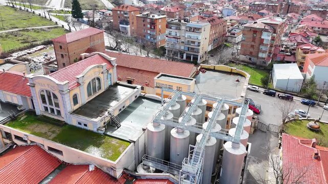 Aerial tracking shot over the historic industrial facades, modern steel fermentation silos, and prominent brick chimney of the Birra Korca factory in Albania