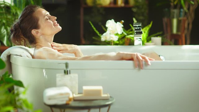 A serene woman with eyes closed gently touches her neck while relaxing in a luxurious bathtub. Surrounded by lush plants & soft natural lighting, she embodies tranquil self-care & indulgent pampering.