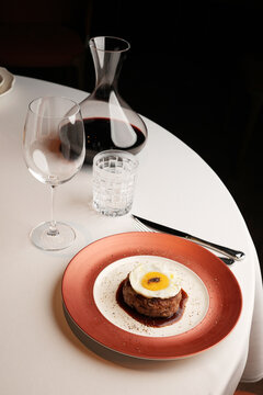 Close-up of a juicy minced beef steak topped with a fried egg on a red rimmed plate, served with red wine in a decanter and empty glasses on a white table. Elegant restaurant main course
