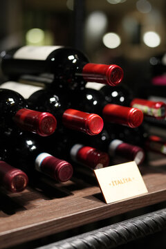 Close-up of red wine bottles with red capsules stacked horizontally on a wooden shelf in a wine cellar or store, featuring a small tag. Moody evening lighting with beautiful bokeh