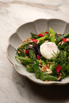 High angle view of a gourmet green salad with fresh asparagus spears, a poached egg, almond flakes, and red flower petals in a ceramic bowl on a marble table. Elegant restaurant appetizer