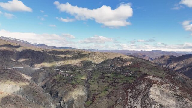 Wide aerial tracking shot panning leftward over a deep mountain chasm, approaching a remote hillside village nestled on green terraced slopes along a winding Balkan highway