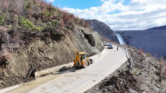 Aerial tracking shot moving forward over a mountain road as a yellow backhoe loader clears landslide debris, with the massive spillway of Moglic&euml; Dam in the background