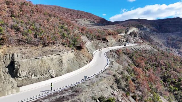Aerial tracking shot raising upward over a curved mountain road, revealing a white minibus traveling along the steep, terraced hillside slopes under a bright blue sky