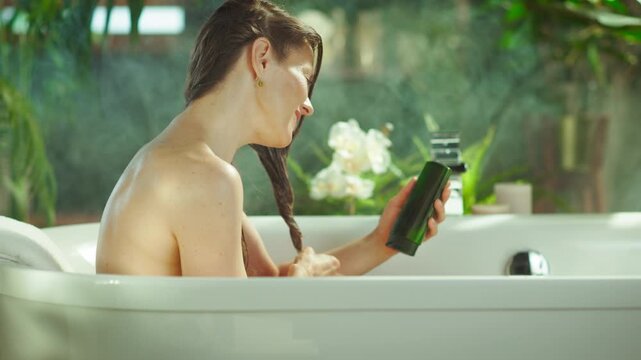 A radiant woman with wet brown hair smiles in a bubbly white bathtub, holding a green hair product & examining her hair. Lush plants & soft light create a serene, spa-like self-care experience.