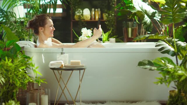 A smiling woman with a bun enjoys a luxurious bubble bath, using a smartphone. Surrounded by lush greenery & candles, capturing self-care, comfort & digital connection in a tranquil setting.