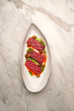 Top view of elegant tuna tataki slices served with citrus wedges, green herb oil, and ponzu sauce on an oval plate over a marble table. Gourmet seafood appetizer in a fine dining restaurant.