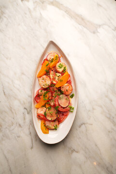 High angle view of a gourmet salad with grilled scallops, orange segments, radish slices, and tomatoes served on an oval ceramic plate over a marble table. Elegant restaurant seafood appetizer