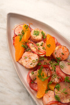 High angle view of a gourmet salad with grilled scallops, orange segments, radish slices, and tomatoes served on an oval ceramic plate over a marble table. Elegant restaurant seafood appetizer