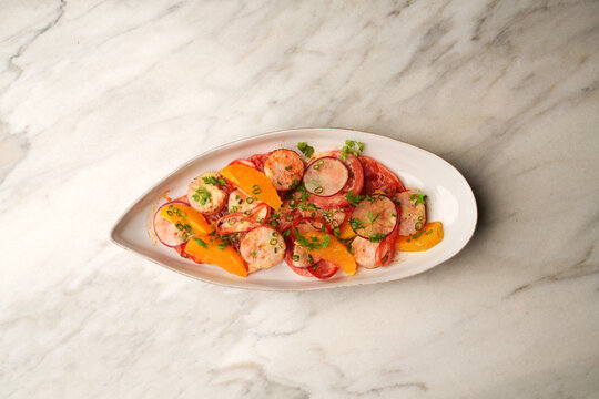 High angle view of a gourmet salad with grilled scallops, orange segments, radish slices, and tomatoes served on an oval ceramic plate over a marble table. Elegant restaurant seafood appetizer