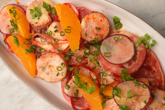 High angle view of a gourmet salad with grilled scallops, orange segments, radish slices, and tomatoes served on an oval ceramic plate over a marble table. Elegant restaurant seafood appetizer