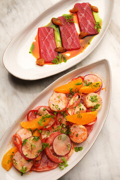 Top view of two elegant seafood dishes, tuna tataki sashimi with sauce and grilled scallop salad with oranges, served on oval plates over a light marble background. High-end restaurant catering