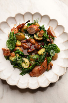 High angle view of gourmet roasted quail pieces served with potatoes, mushrooms, fresh greens, and rich brown gravy in a ceramic plate on a marble table. Elegant restaurant poultry dish