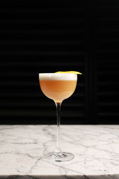 Classic sour cocktail with thick white foam and a yellow lemon twist garnish, served in a coupe glass on a marble table against a dark background. Fine dining bar drink